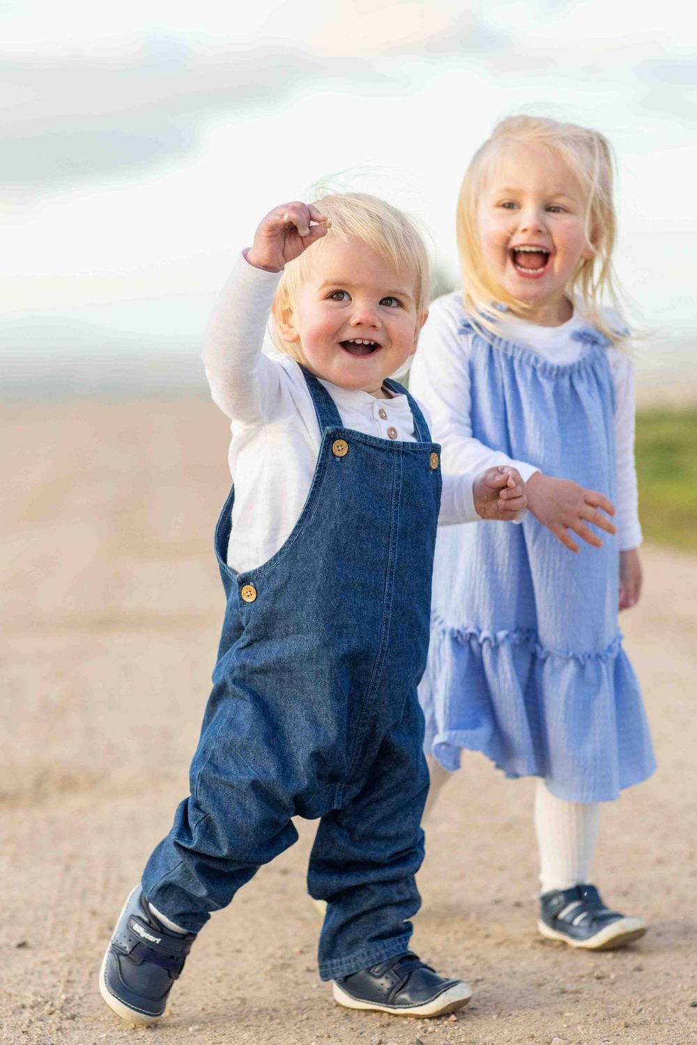 Two toddlers playing together on a sandy beach, one wearing denim overalls and one in a blue dress.