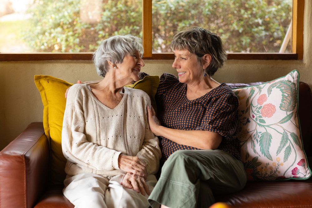 Two people share a warm moment while sitting together on a couch in a sunlit living room.