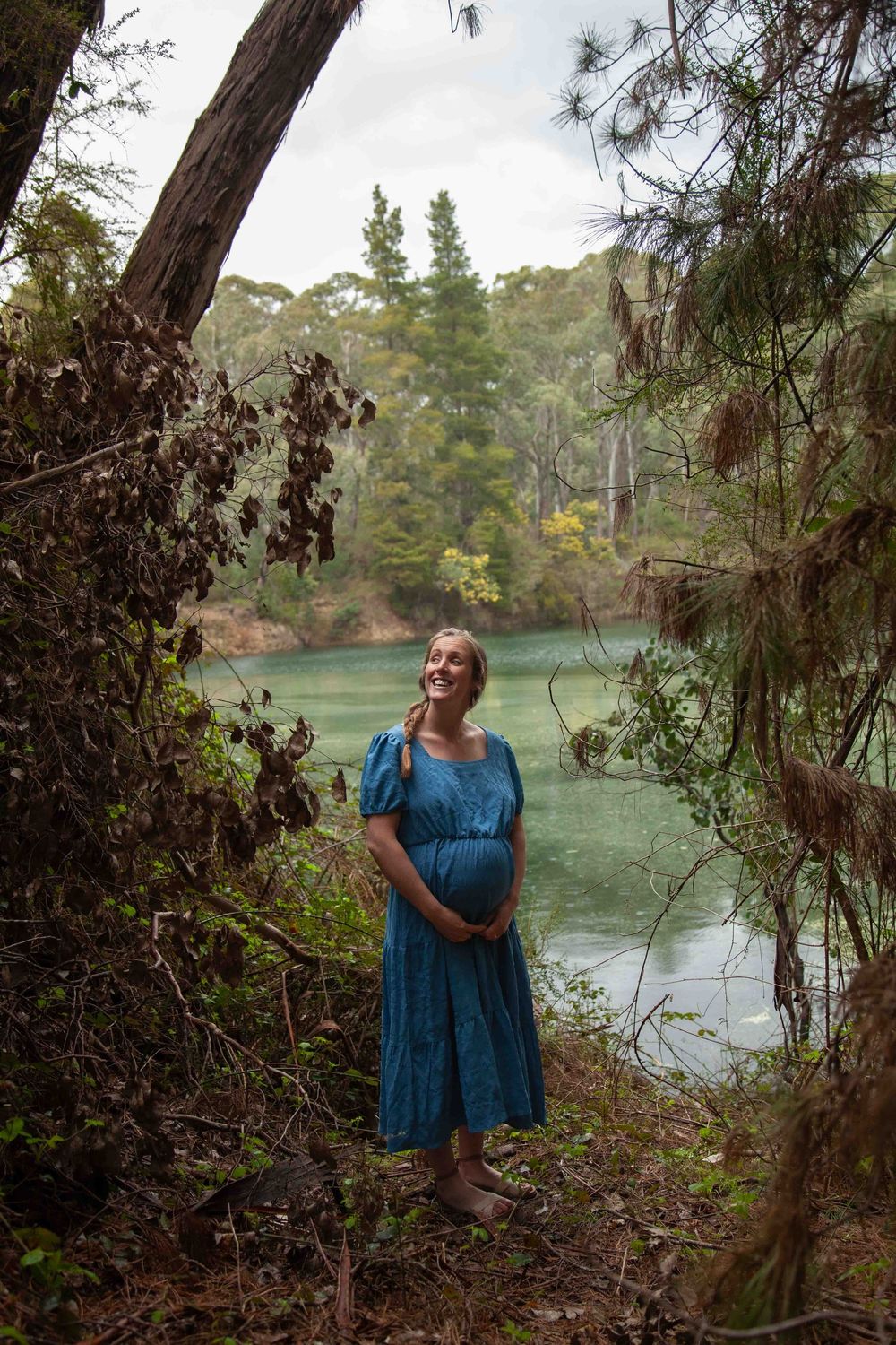 A figure in a blue dress stands among trees beside a tranquil lake on a misty afternoon.