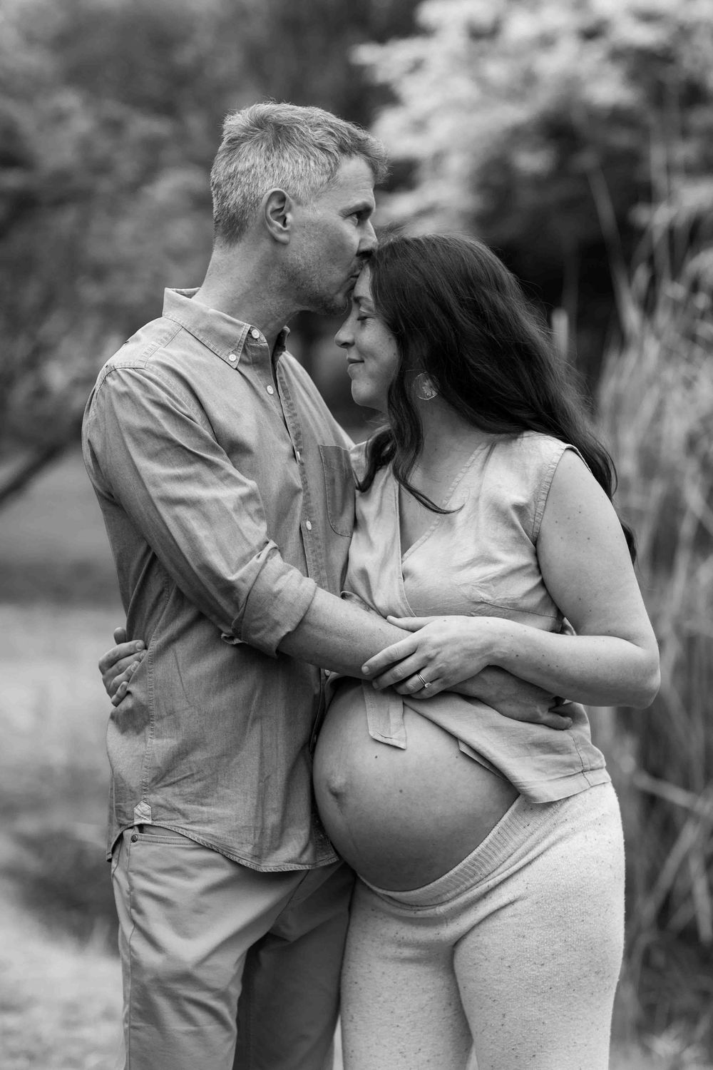 A tender black and white maternity photo of a couple embracing outdoors in a natural setting.