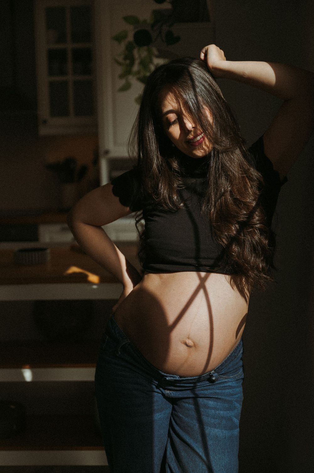 Pregnant woman in black crop top and jeans posing in sunlight, showing baby bump indoors.