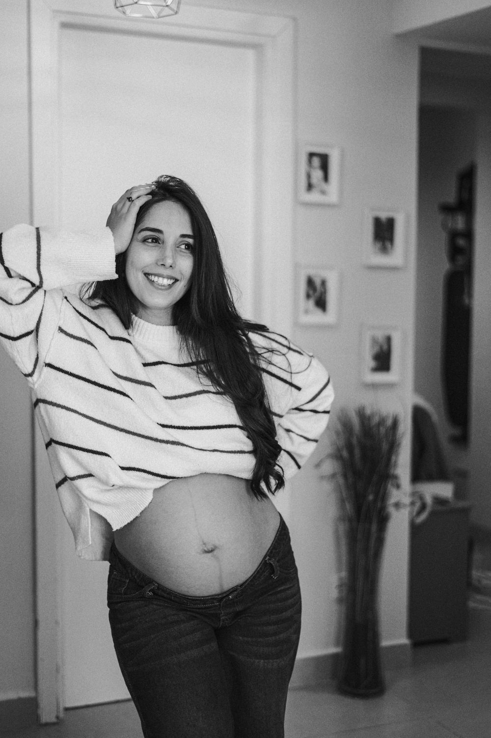 Smiling pregnant woman in striped sweater and jeans posing indoors, showing baby bump in black and white photo.