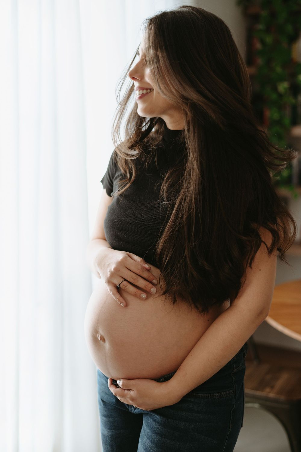 Smiling pregnant woman in black crop top and jeans cradling her baby bump while standing by a bright window.