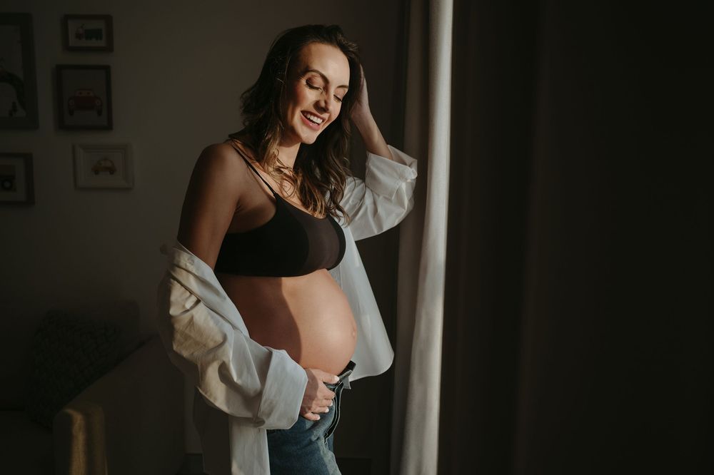 Smiling pregnant woman in black bralette and jeans poses by window, showing baby bump in moody indoor maternity photo.