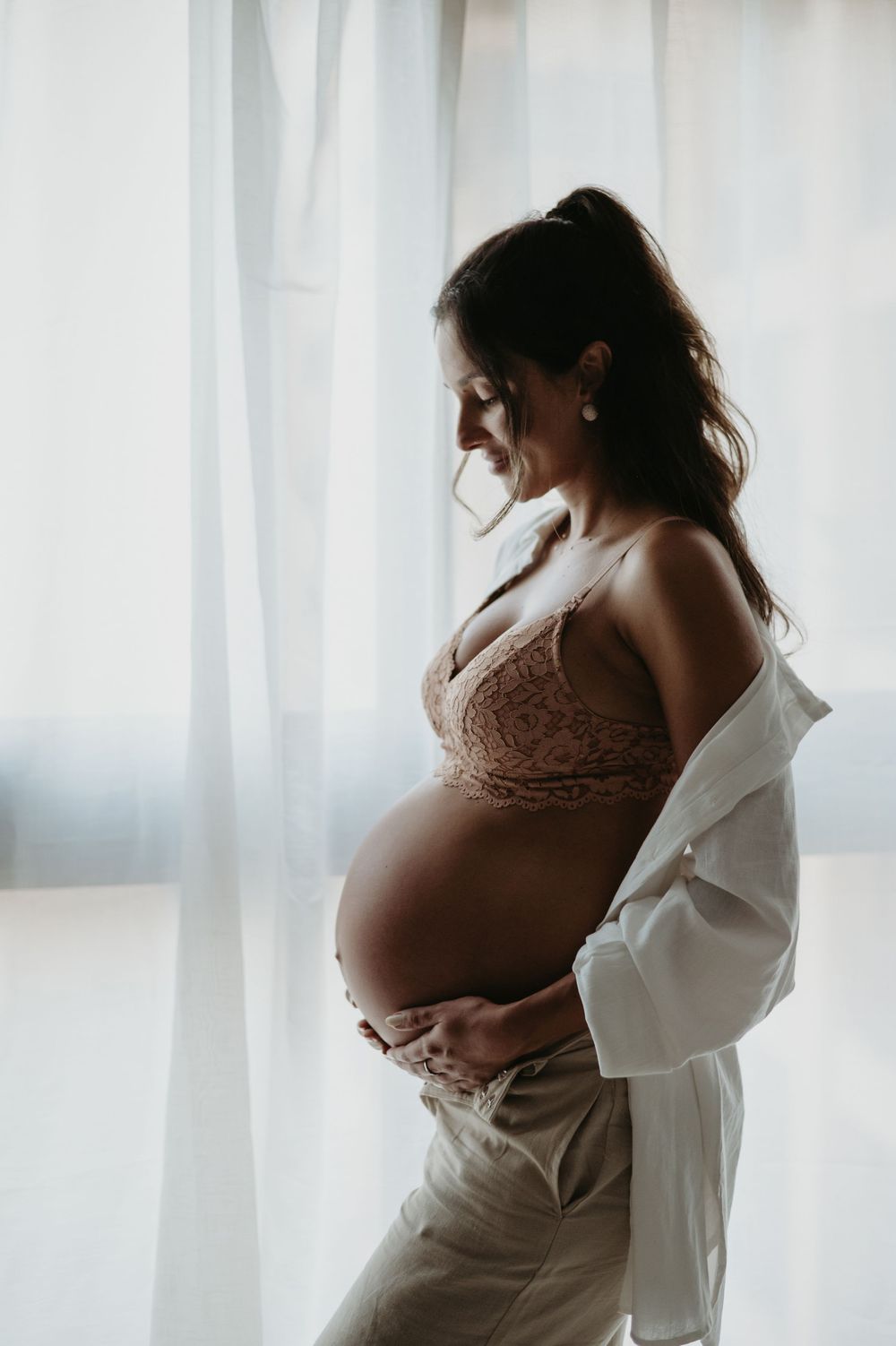 Pregnant woman in lace bralette holding baby bump by sheer curtained window in soft natural light.