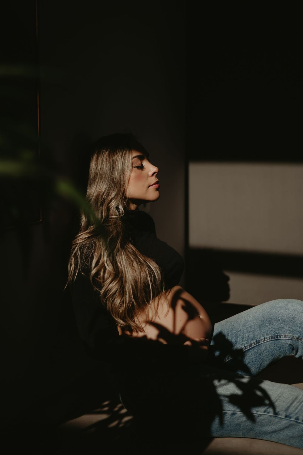 Woman with long blonde hair in black turtleneck and jeans sits peacefully in dramatic shadow and light.