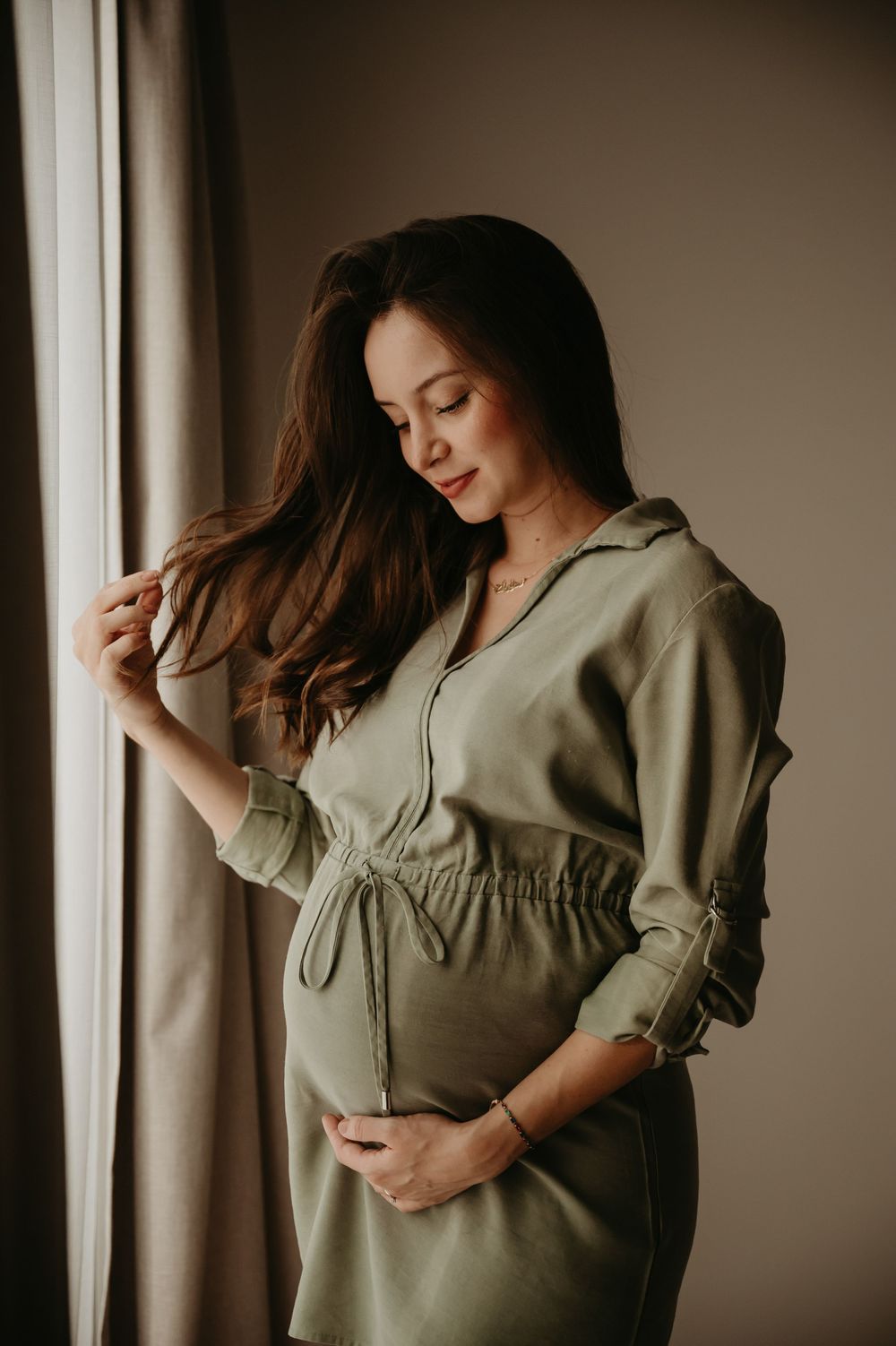 Pregnant woman in olive green dress standing by window, cradling baby bump with one hand, touching hair.