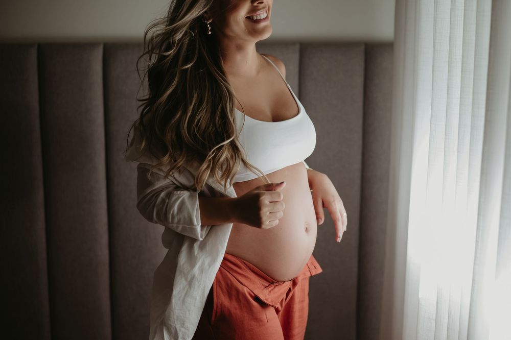 Smiling pregnant woman in white crop top and red pants cradling her baby bump near a window.