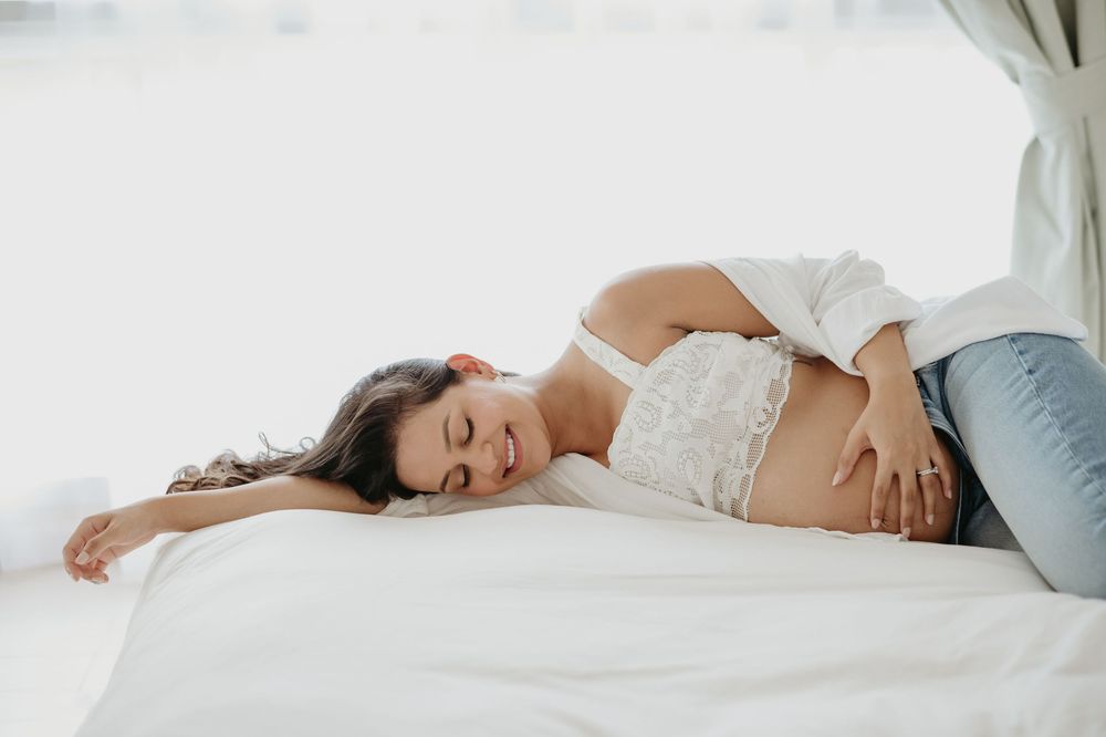 Pregnant woman resting on white bed, cradling baby bump, wearing white lace top and jeans.