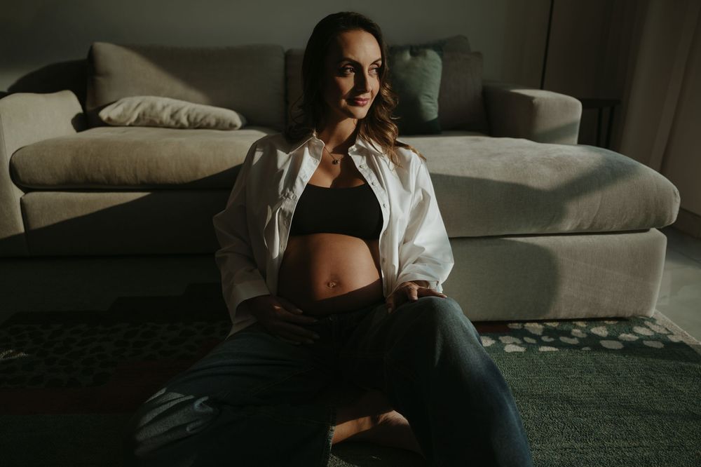 Pregnant woman sitting on floor in white shirt and black crop top, cradling baby bump in cozy living room.