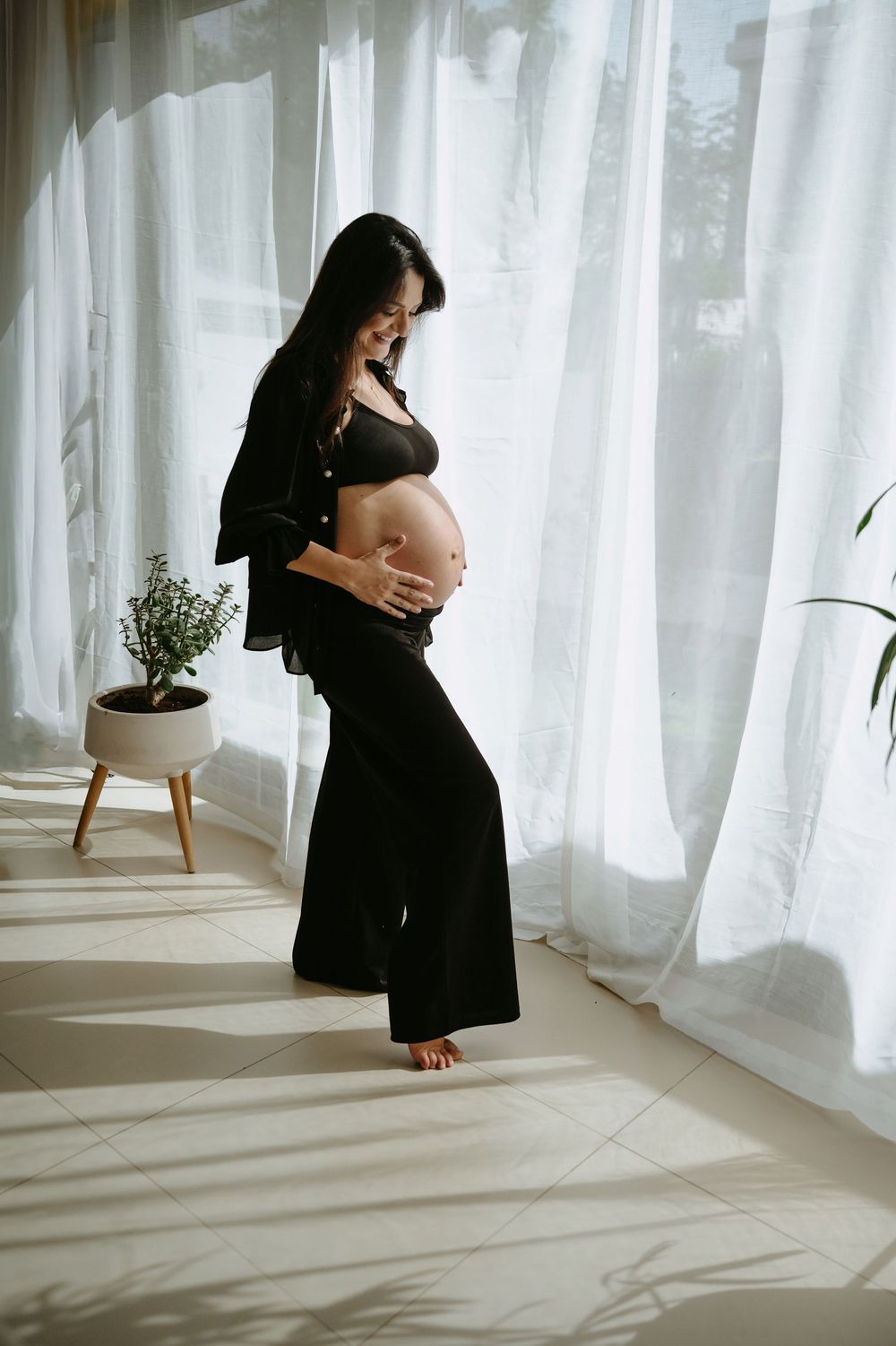 Pregnant woman in black outfit cradling baby bump, standing by sheer curtains in sunlit room.
