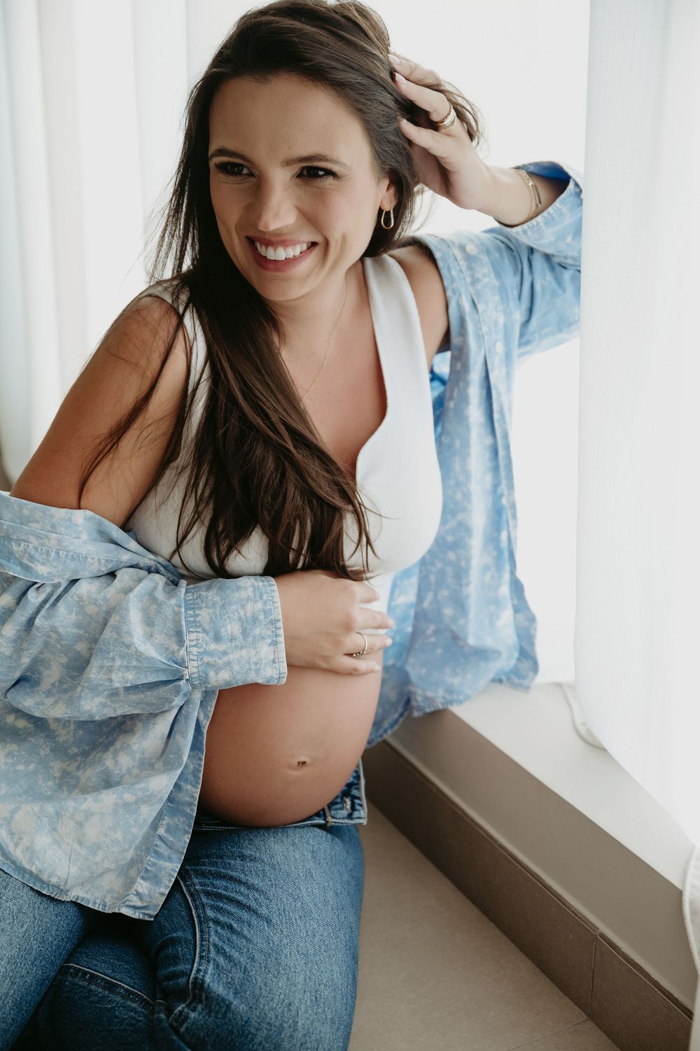 Smiling pregnant woman in blue shirt and jeans cradling her baby bump while sitting by a bright window.