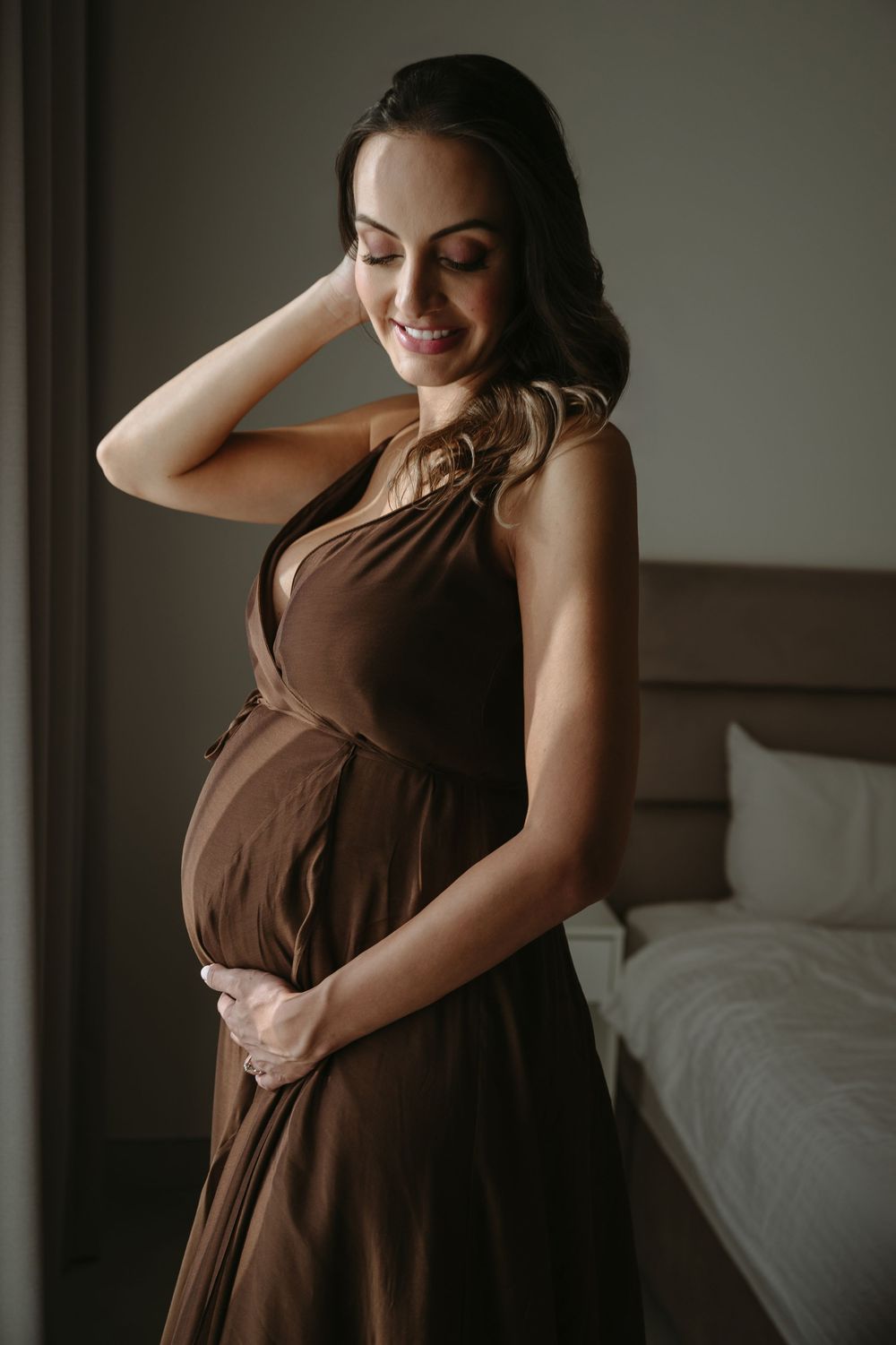 Smiling pregnant woman in brown halter dress cradling baby bump in softly lit bedroom.