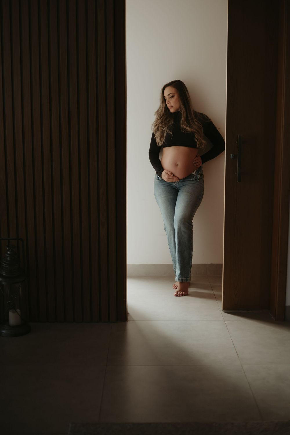 Pregnant woman in black crop top and jeans cradling baby bump while leaning against wall in modern home.