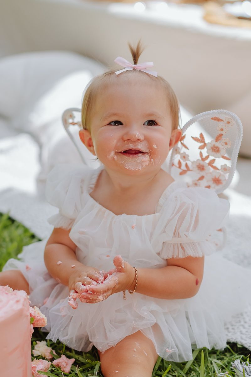 one year old girl smiling wearing angel wings for a cake smash photo session