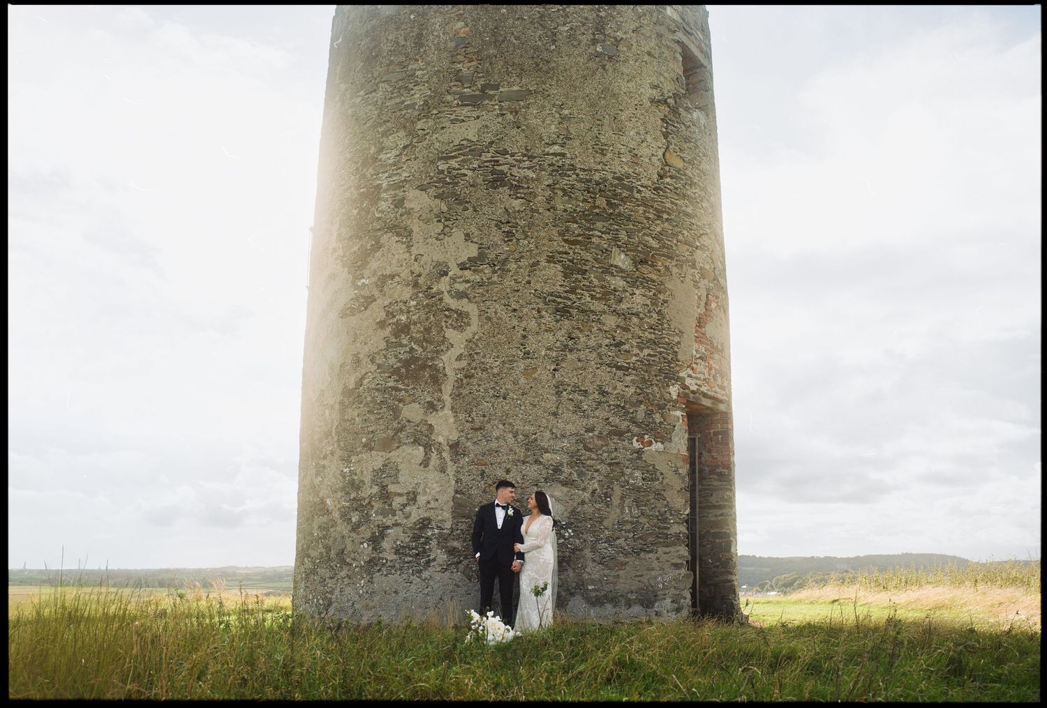 Wedding portraits captured against a tall stone lighthouse tower under cloudy skies.