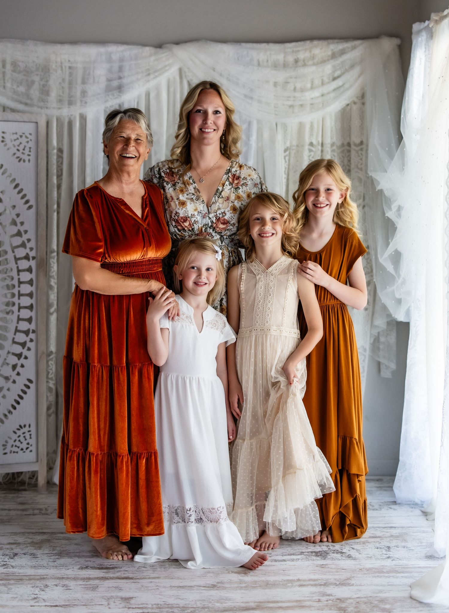 A group in white dresses and rust-colored outfits pose together in front of a white lace backdrop.
