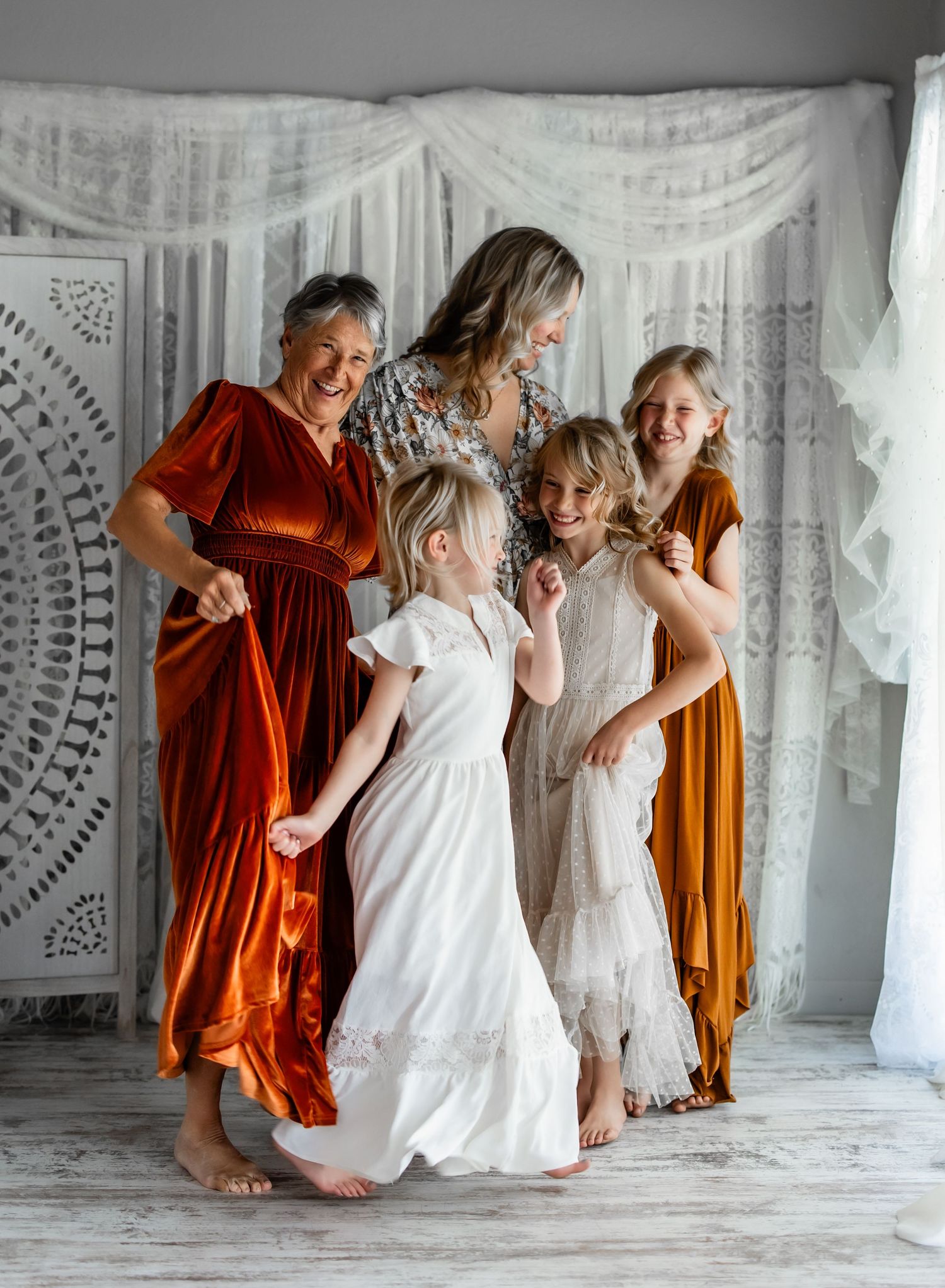 A group in rust-colored and white dresses dance and twirl together in a light-filled room with lace curtains.