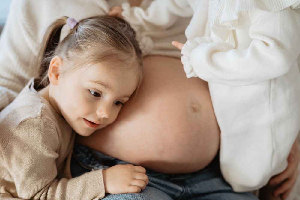 A pregnant woman lies on white bedding while a child rests their head on her belly.