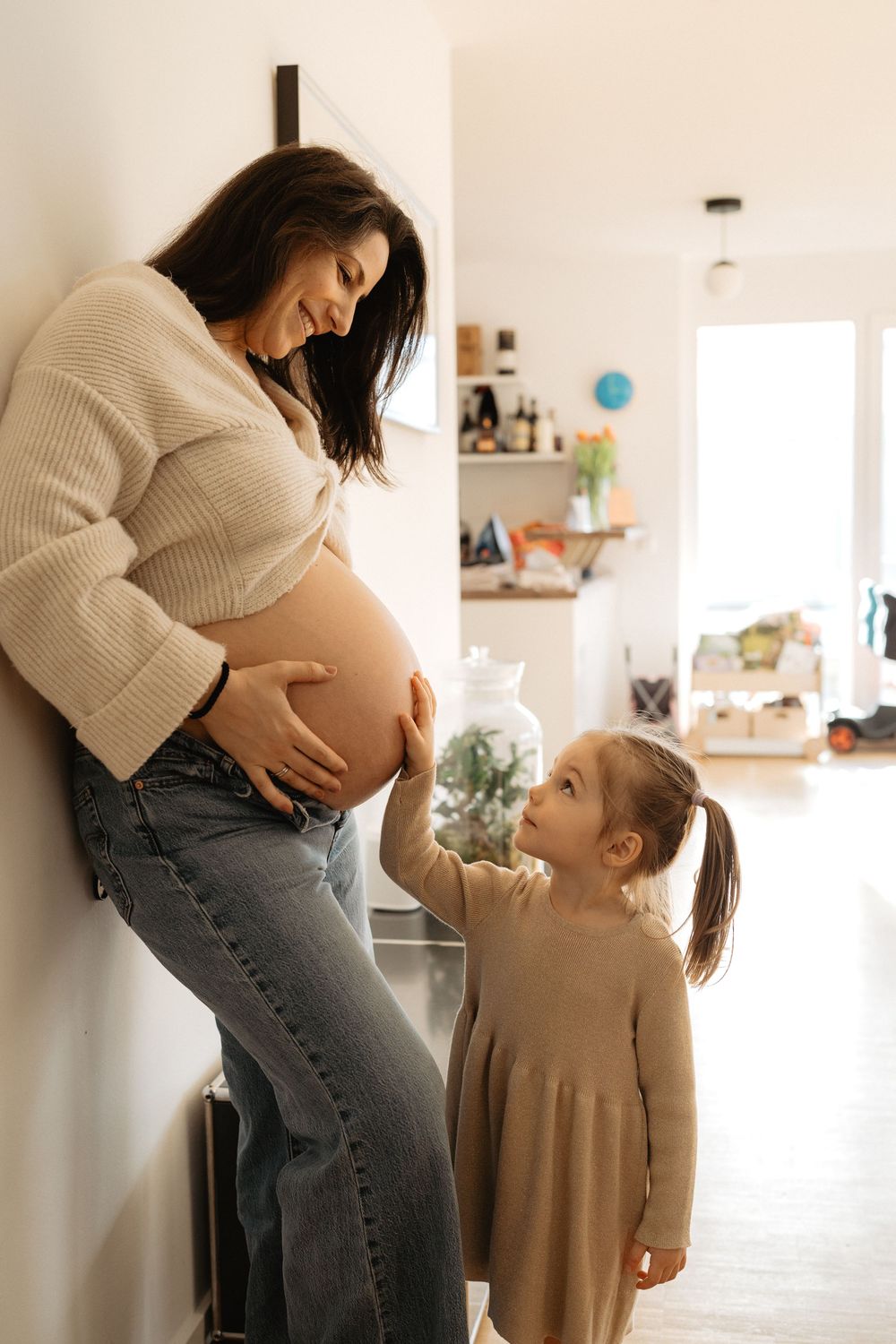 A pregnant woman in a cream sweater and jeans shares tender moments with a young child in a bright home setting.
