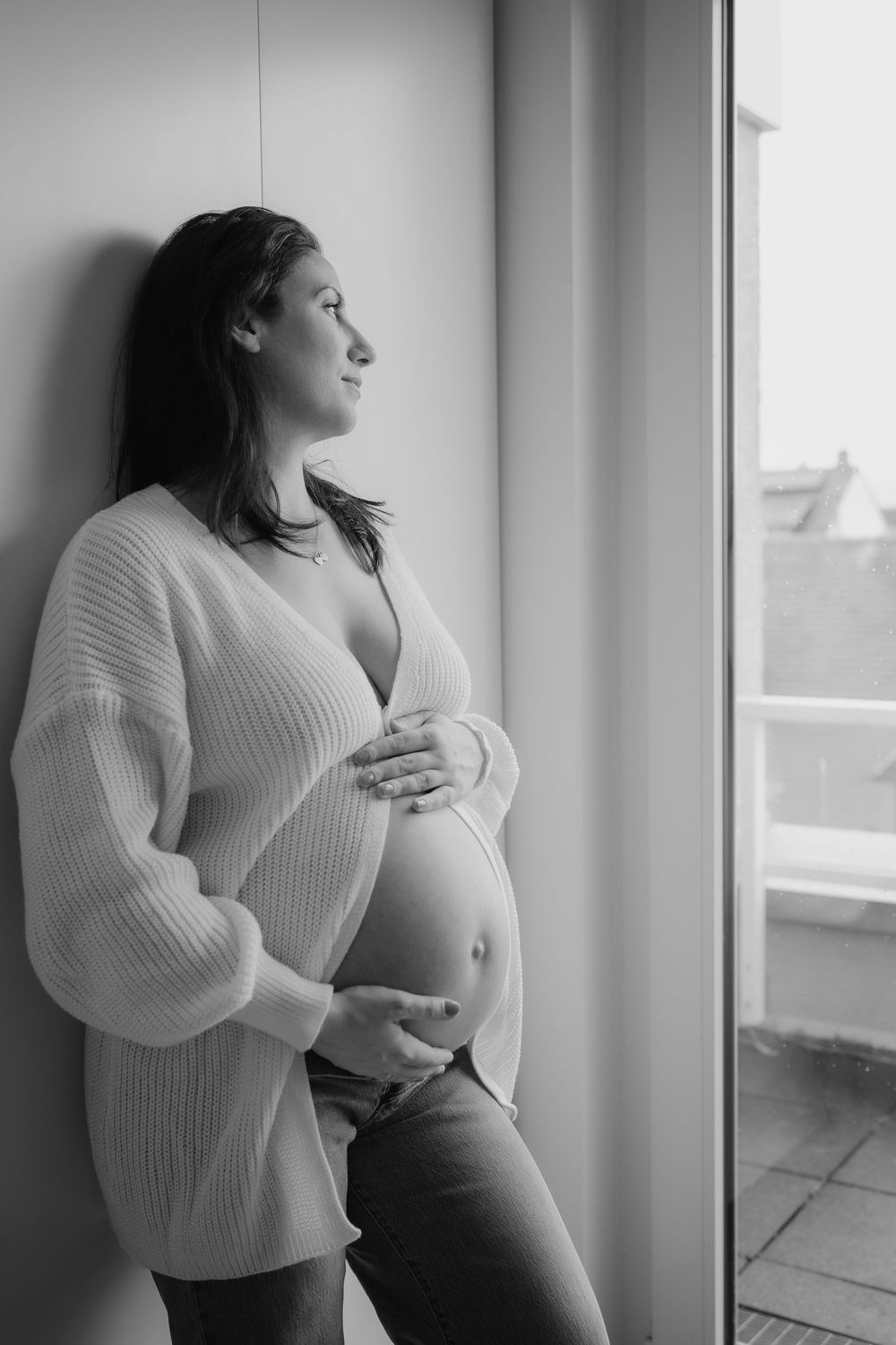Black and white maternity portraits showing a woman in a cozy sweater by a window.
