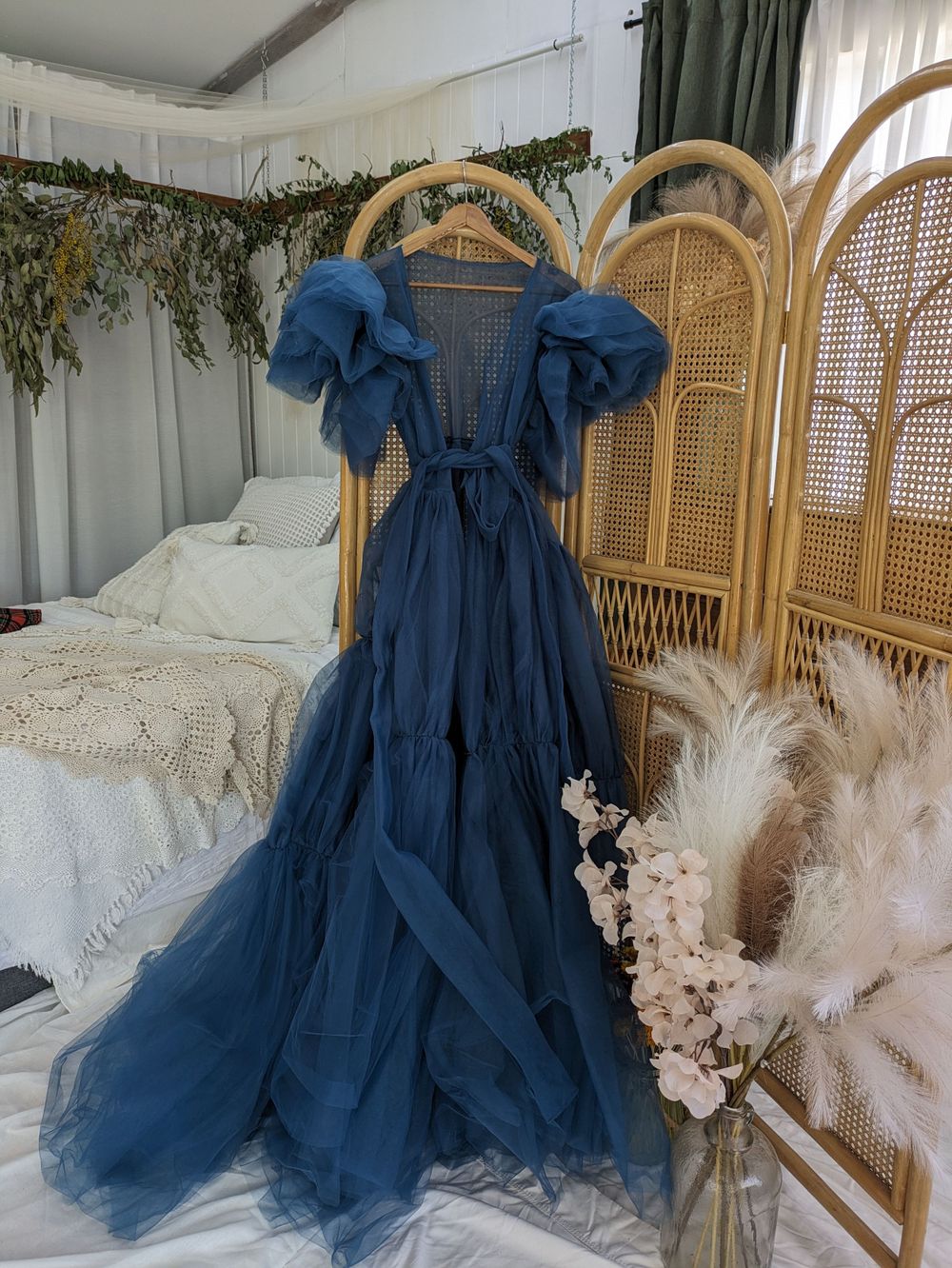 Ethereal blue tulle gown with puff sleeves displayed against rattan screen with dried flowers and white bedding.