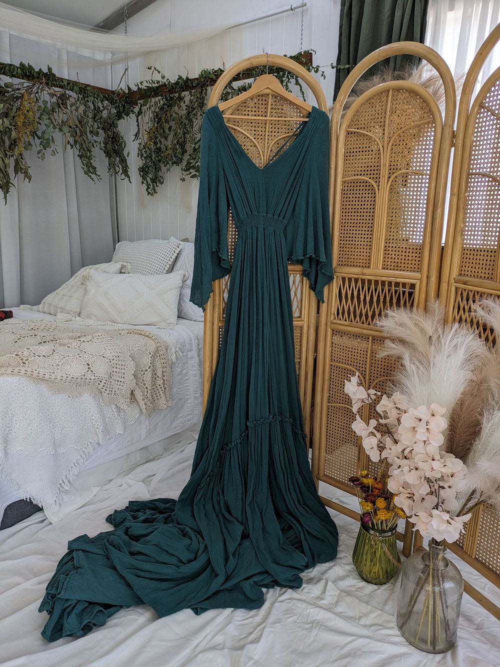Emerald green flowing dress with long sleeves displayed against rattan divider and white bedding.