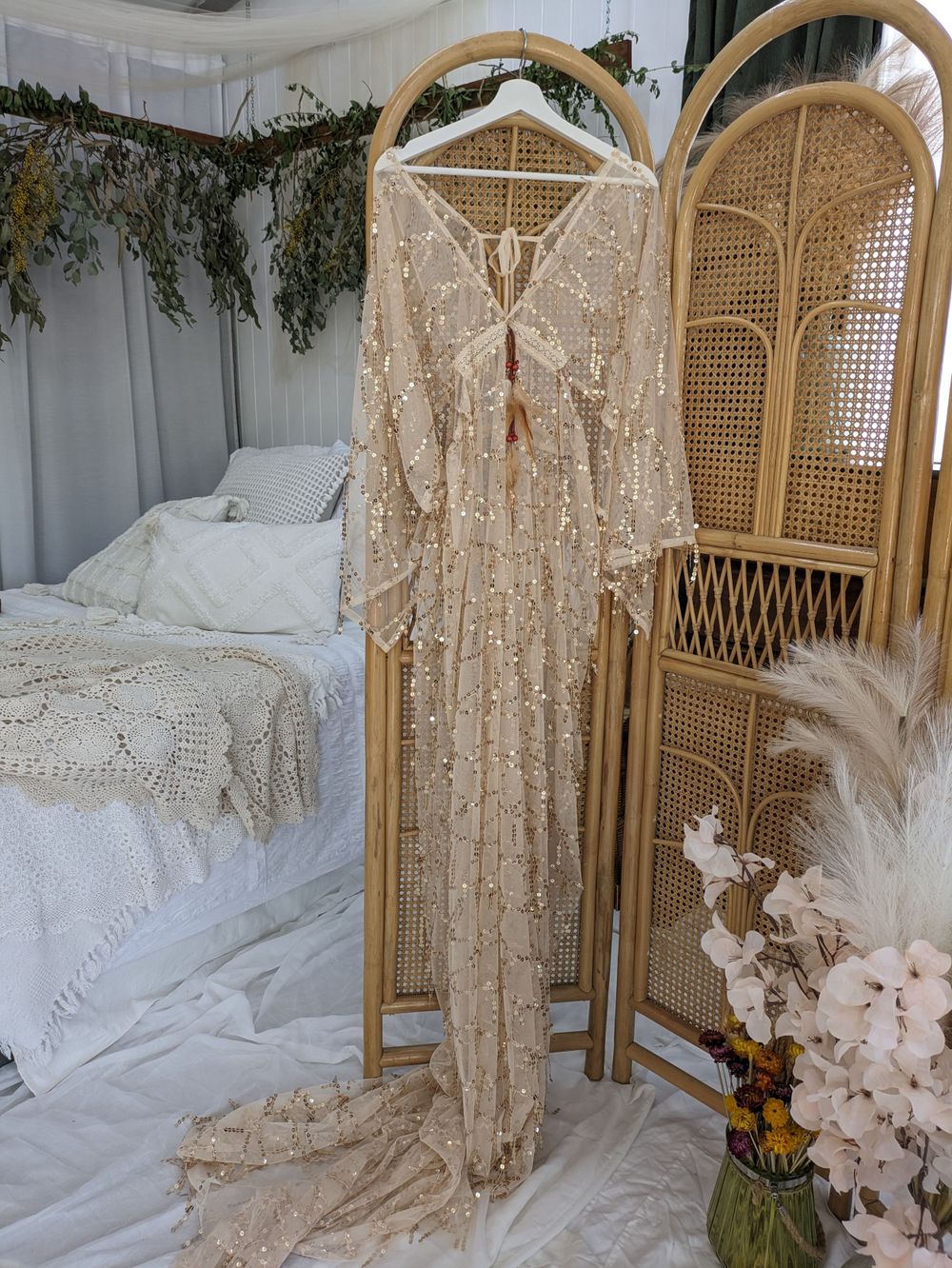 Lace wedding dress hanging on display with rattan screen and floral arrangements.
