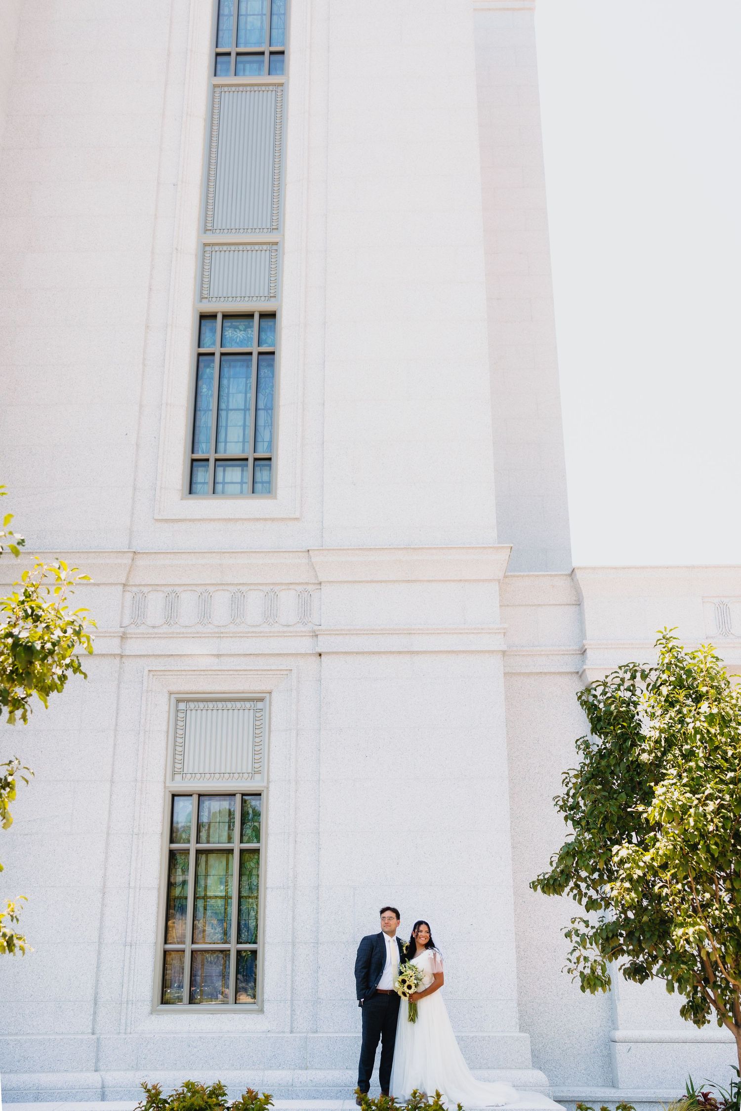A bride in a white gown holding a bouquet and a groom in a dark suit pose beside a tall white Lindon Utah temple with narrow windows.