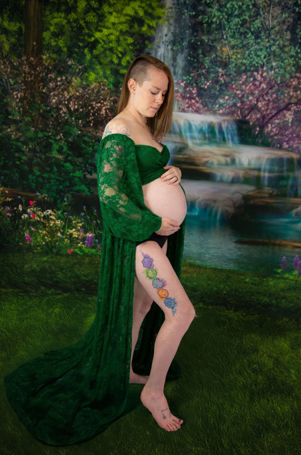 Maternity photo of an expectant mother in an emerald green lace gown standing by a waterfall in a dreamy garden setting.