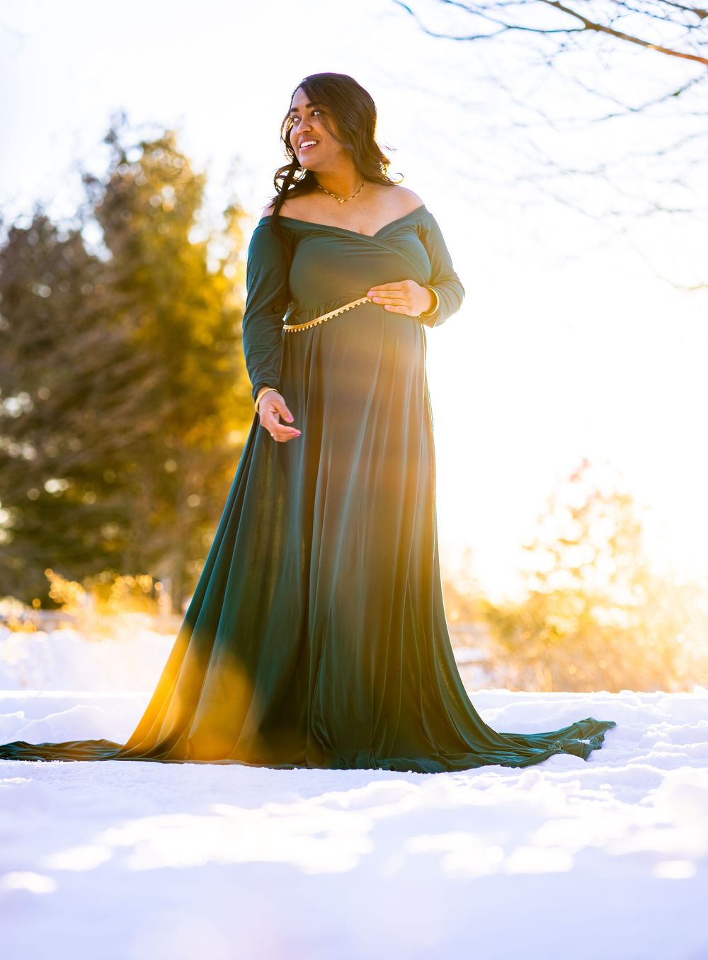 A flowing dark green dress billows in the winter sunlight against a snowy landscape with golden sunset lighting.