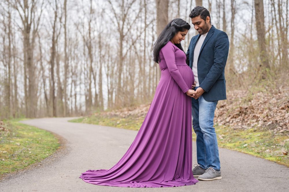 A couple stands on a wooded path with the expectant mother wearing a flowing purple maternity gown.