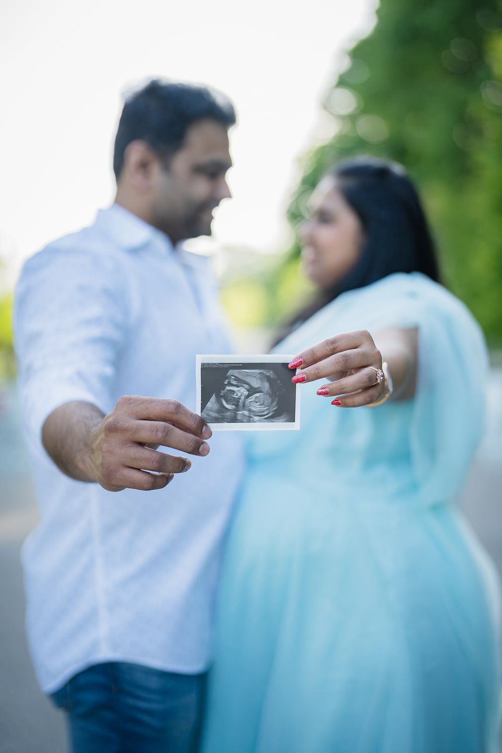 Couple in light blue attire sharing an ultrasound photo outdoors during a pregnancy photoshoot.