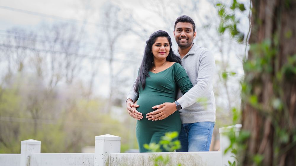 A couple poses by a white fence during a maternity photoshoot in casual attire.