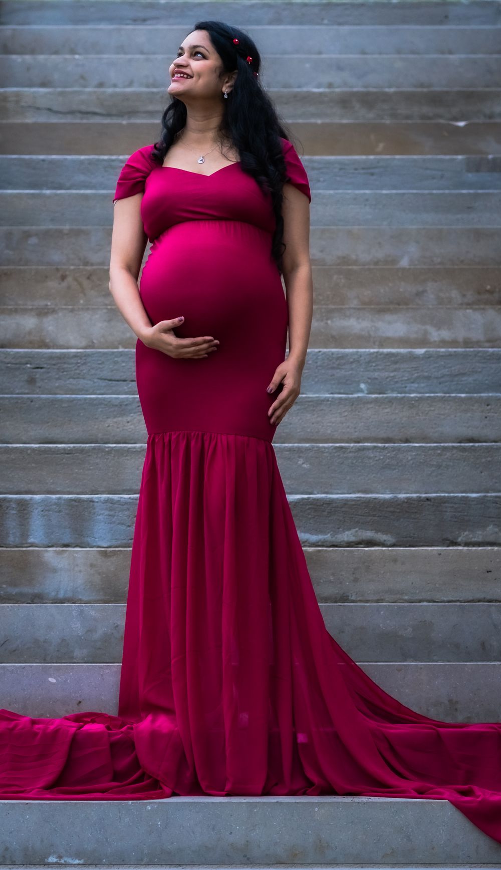 A flowing burgundy maternity gown with long train against a rustic wooden wall backdrop.