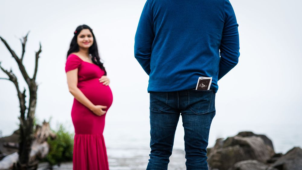 A couple in navy blue outfits stands together outdoors during a maternity photo session.
