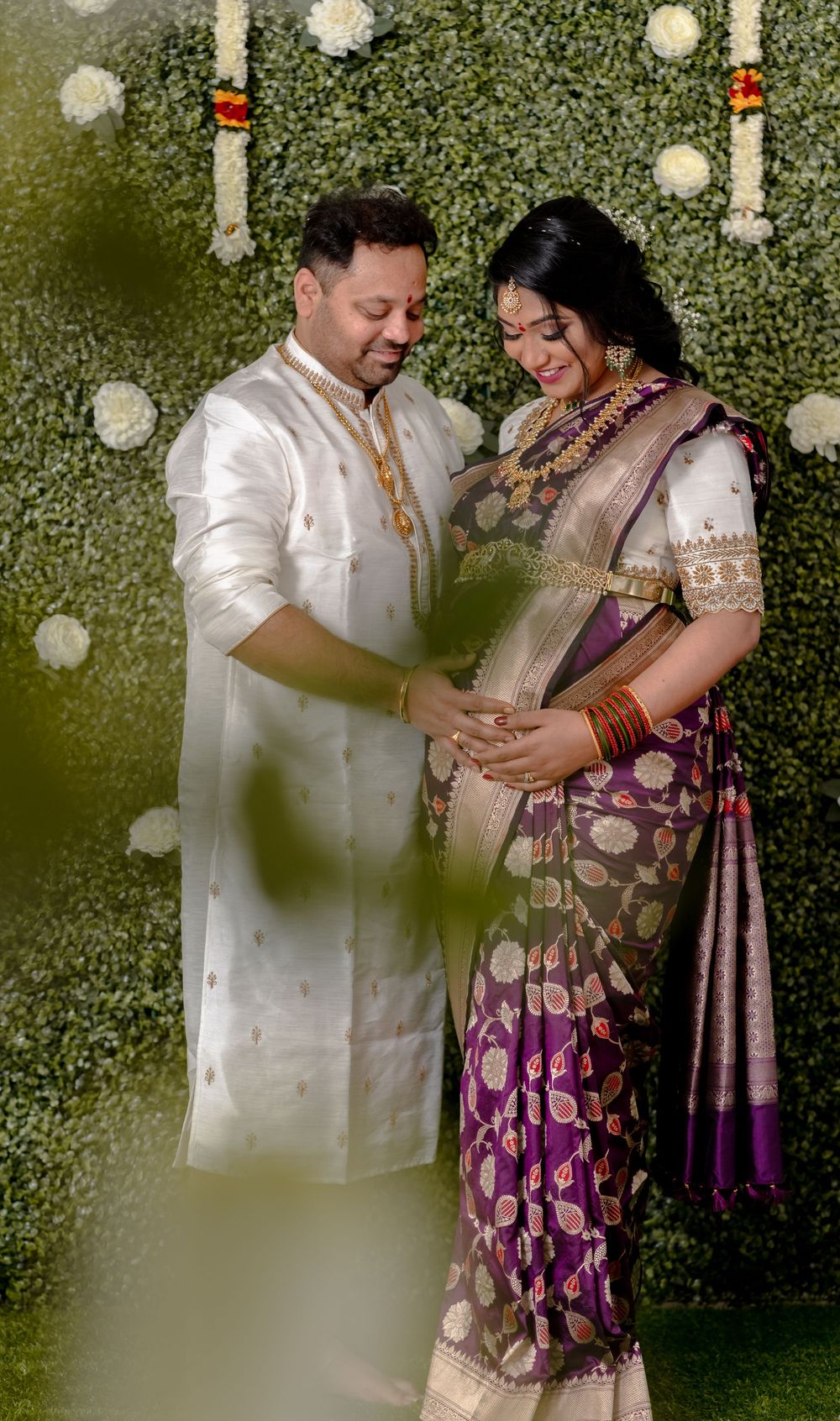 A couple in traditional Indian attire share an intimate moment against a floral backdrop during a maternity photoshoot.