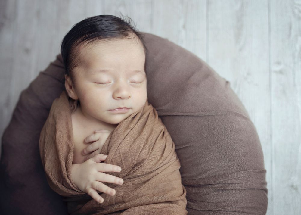 Sleeping newborn wrapped in brown fabric on a round cushion against white backdrop.