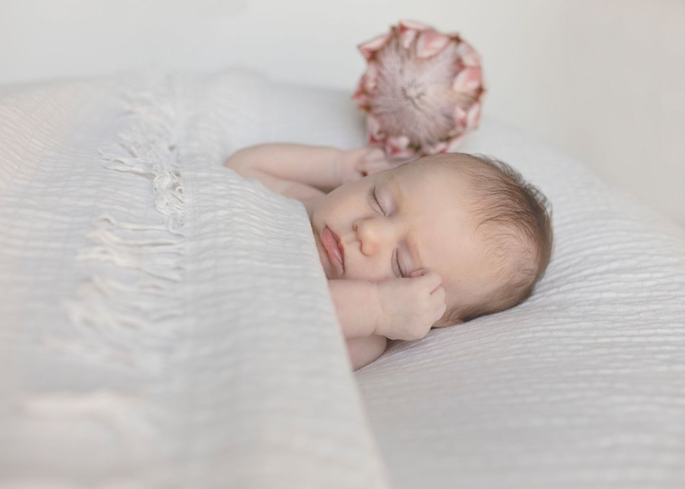 Sleeping newborn with pink flower accessory on white textured bedding.