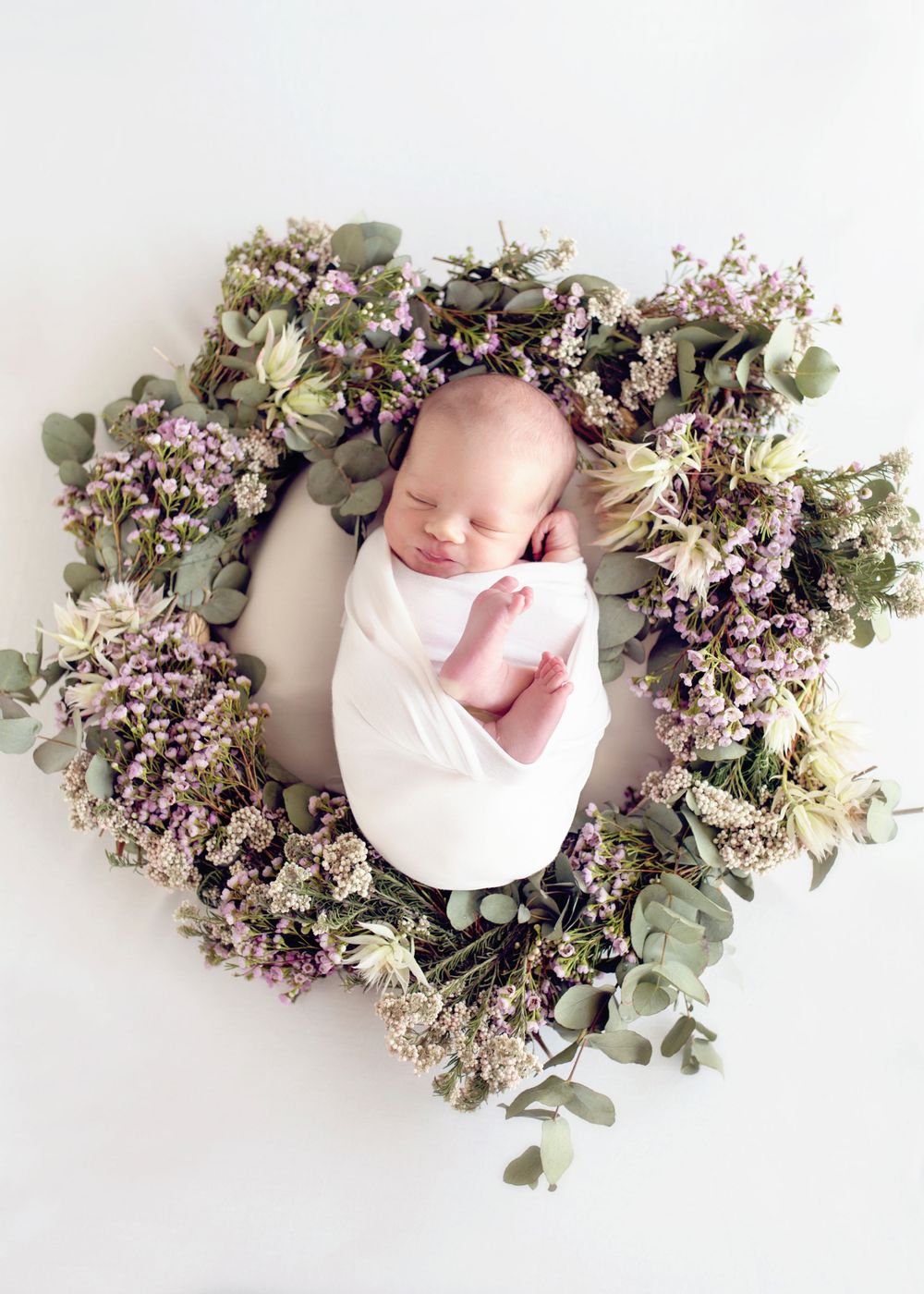 Newborn baby wrapped in white, nestled in a wreath of dried flowers and eucalyptus.