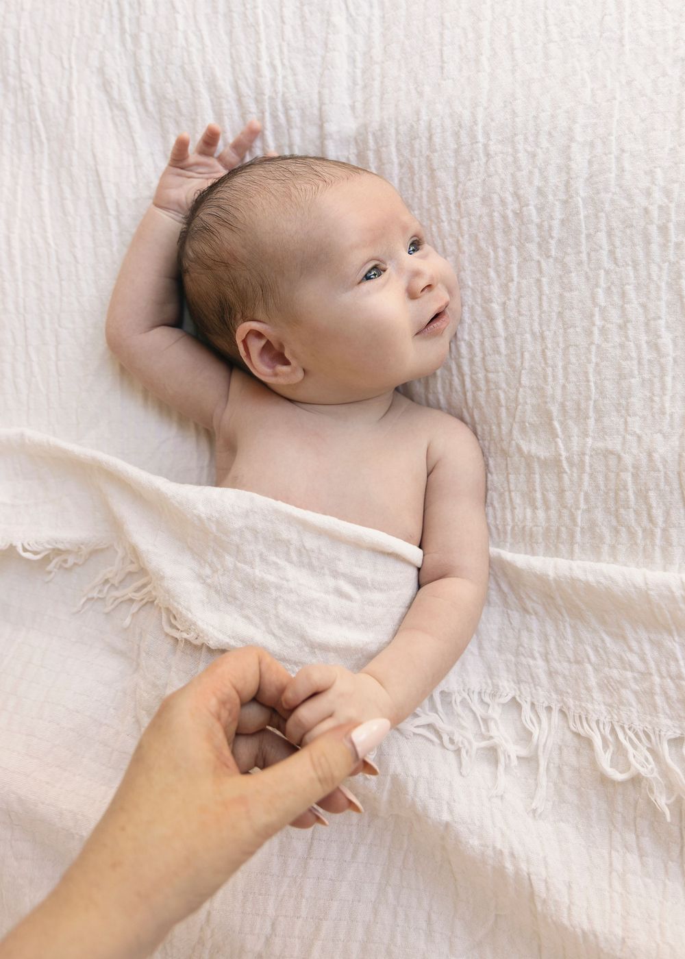 Baby lying on white blanket gazes up with curious expression while adult hand reaches to touch.