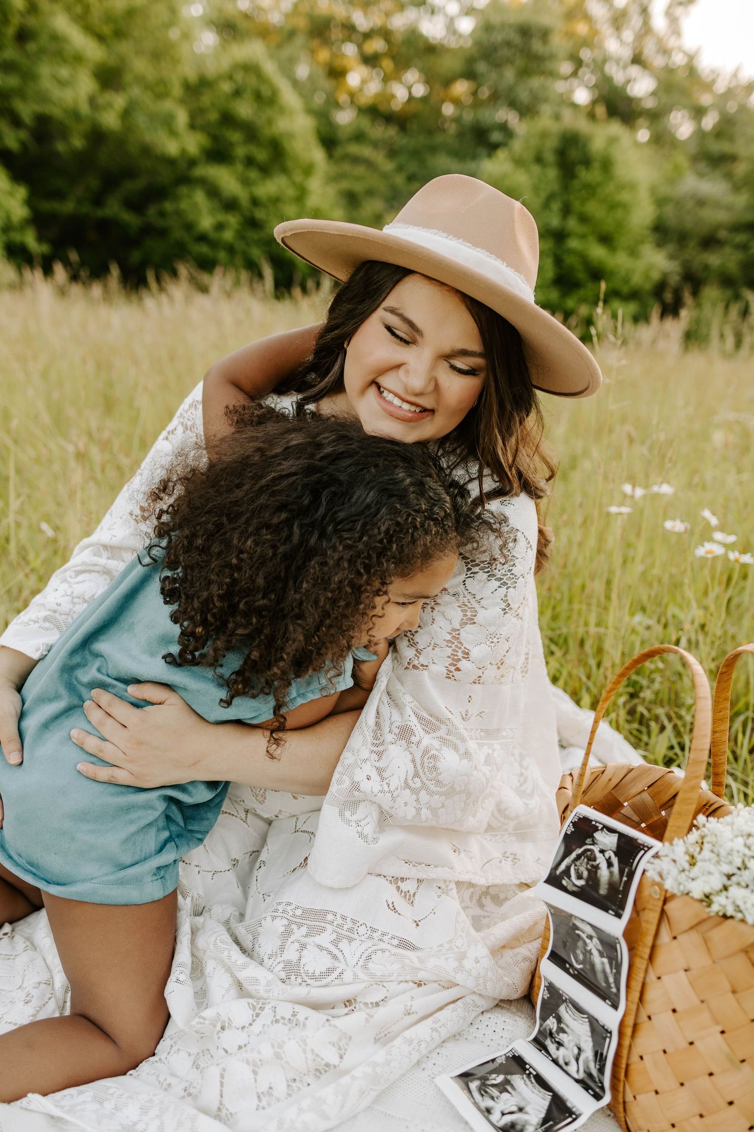 Person in hat embracing child during outdoor photoshoot with ultrasound photos.