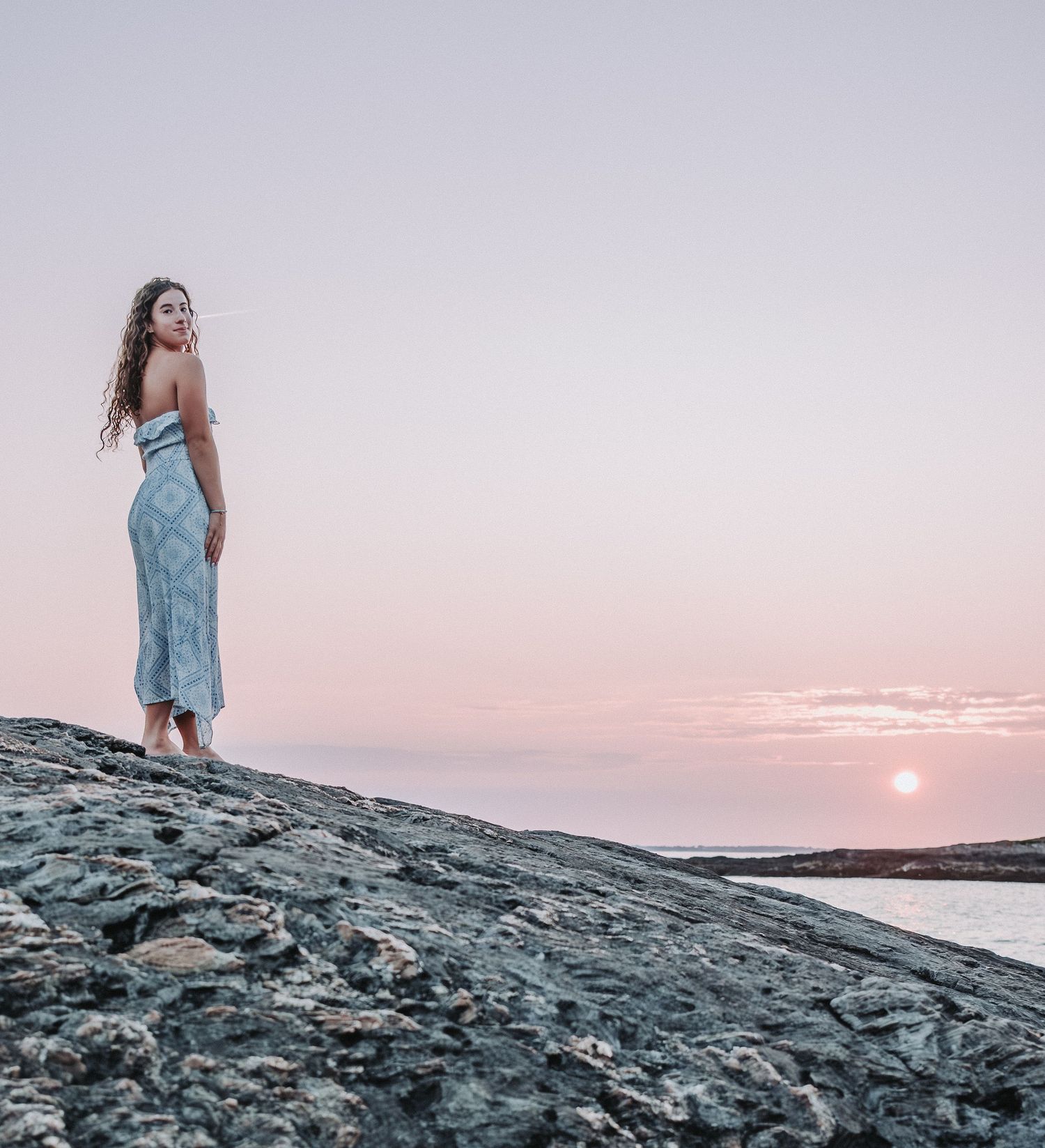 A high school senior stands on rocky coastal terrain with pink and orange sky. during a sunset photo session at Hermit Island.