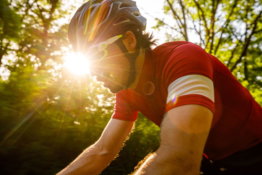 Cyclist in red jersey and helmet rides through sunlit forest, with golden sun rays shining through green trees.