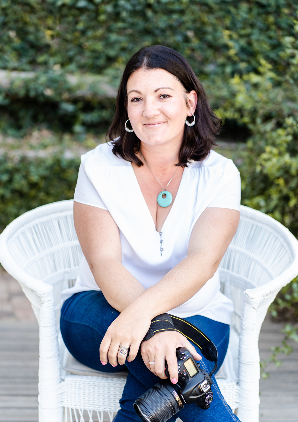 A photographer in white blouse and blue jeans sits casually outdoors with a professional camera in natural lighting.