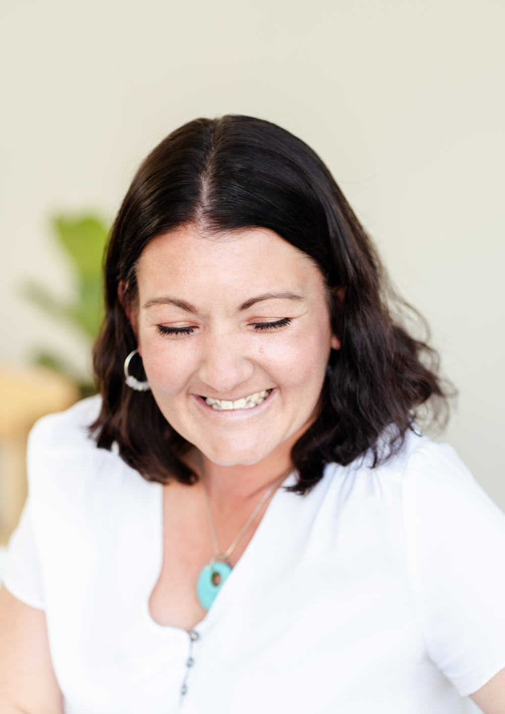 Woman in white t-shirt smiling and looking down while sitting at a table with natural lighting.