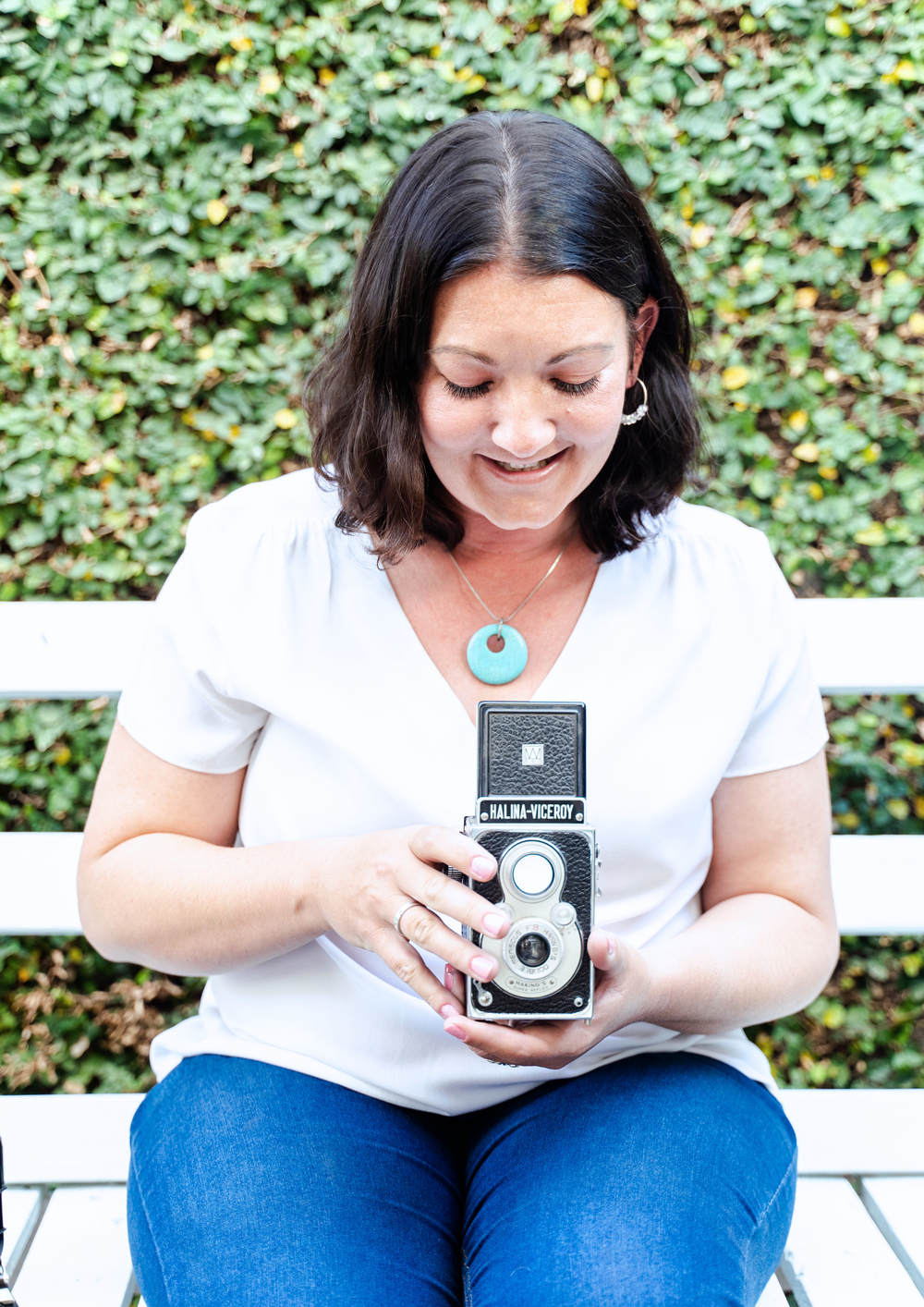 A photographer in a white shirt and jeans sits on a bench holding a vintage twin lens reflex camera against a leafy background.