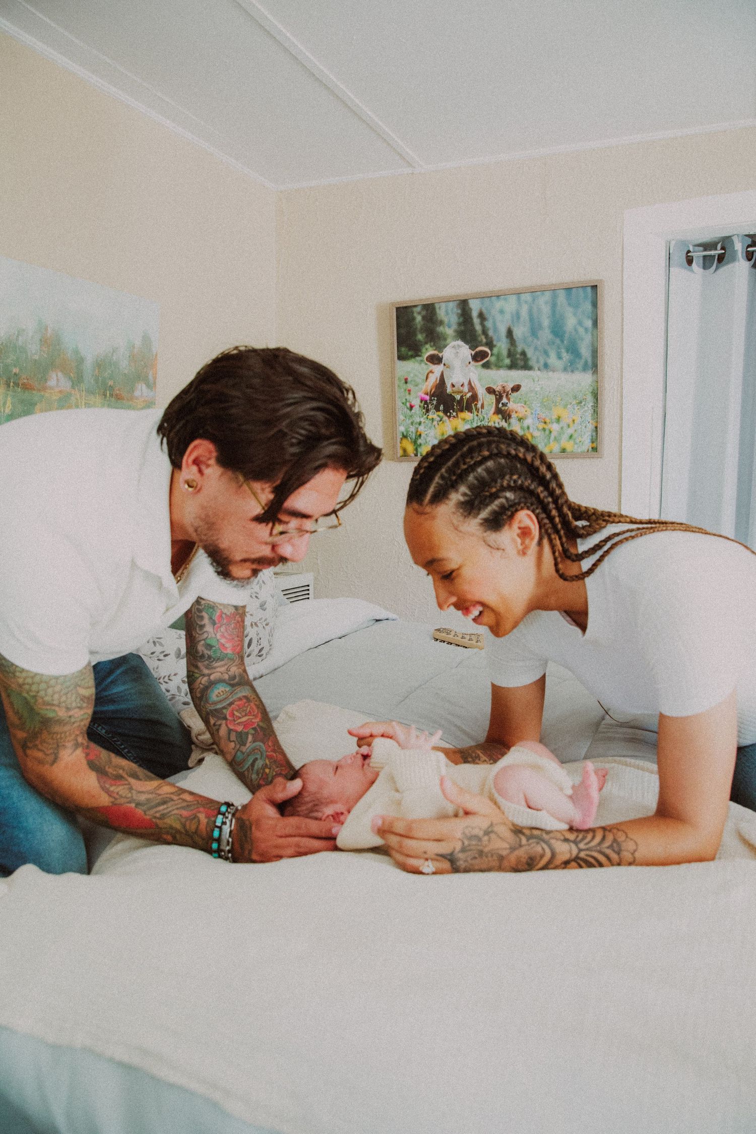 Two tattooed parents lovingly gaze at their newborn baby lying on a white bed in a cozy room.