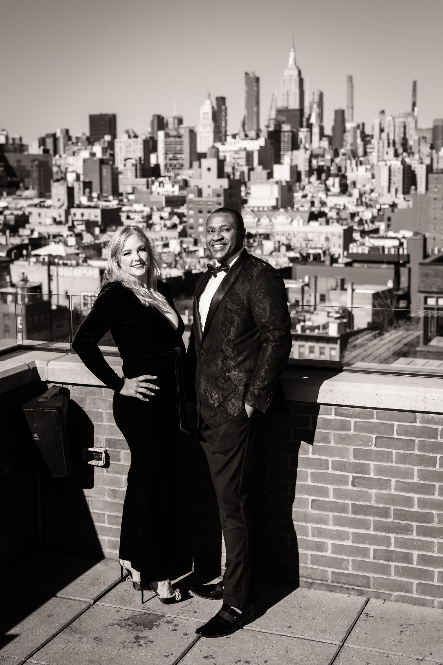 A black and white cityscape photo of a couple in formal attire standing on a rooftop with the New York City skyline behind them.
