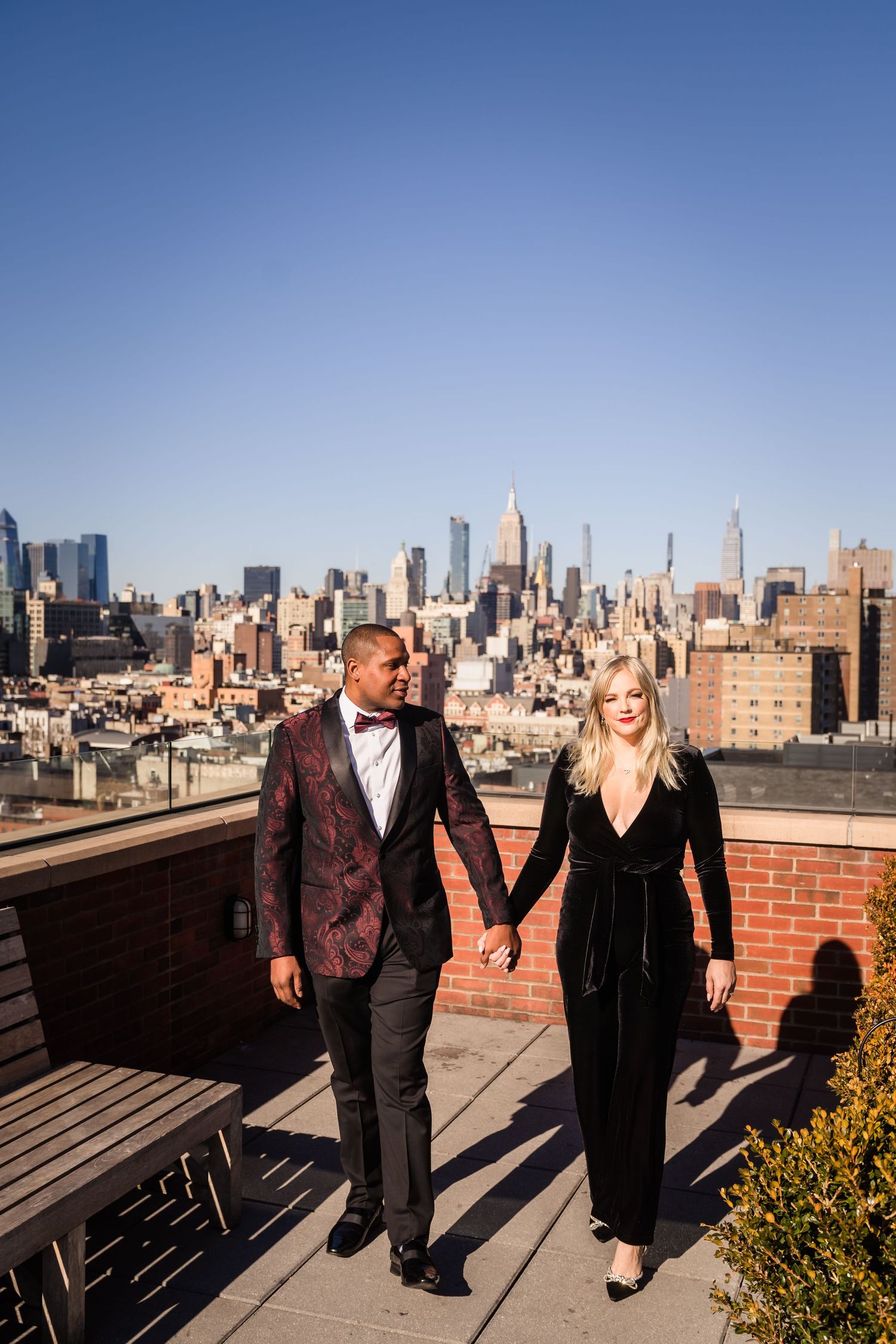 Couple walking hand in hand on a rooftop terrace with Manhattan skyline backdrop on a sunny day.